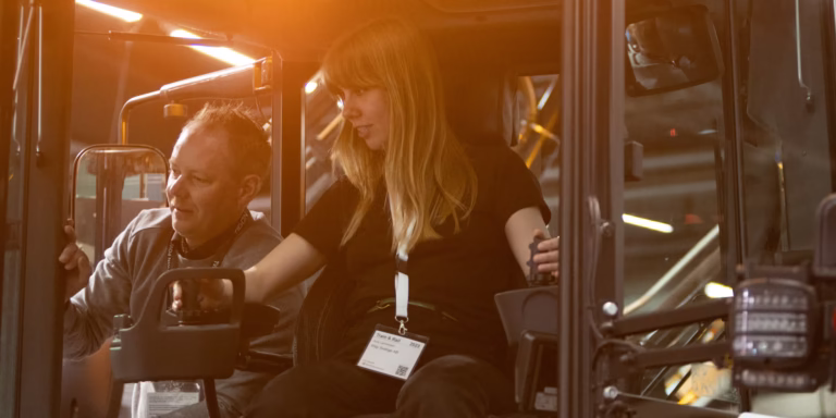 Two people are sitting in the cab of a work vehicle; a man is showing something to a woman who is holding the controls.